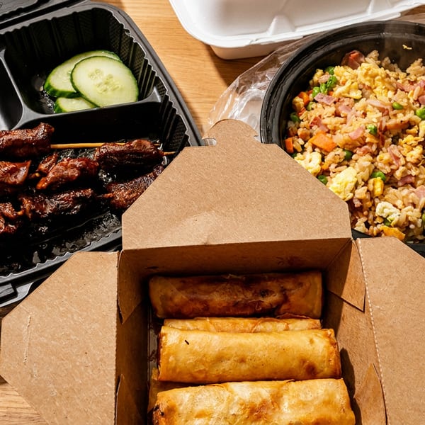 Takeout containers filled with fried rice, spring rolls, and meat skewers with cucumber slices.