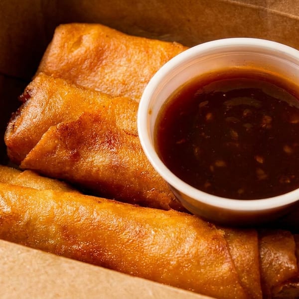 Golden-brown spring rolls and a cup of dipping sauce in a brown takeout box.