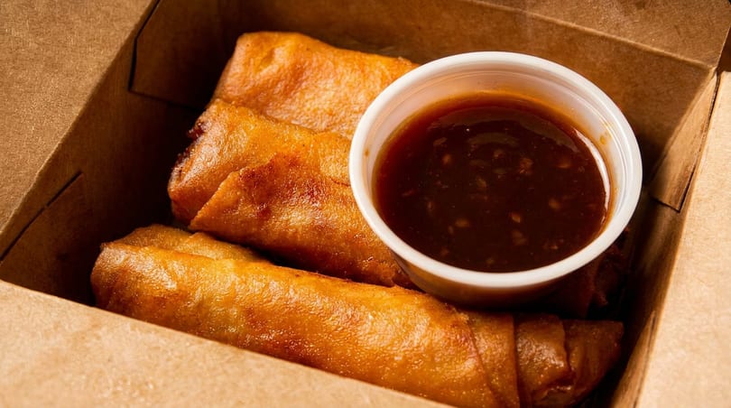 Golden-brown spring rolls and a cup of dipping sauce in a brown takeout box.